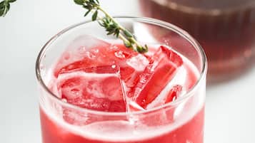 A glass of red beverage with ice cubes and a sprig of thyme.