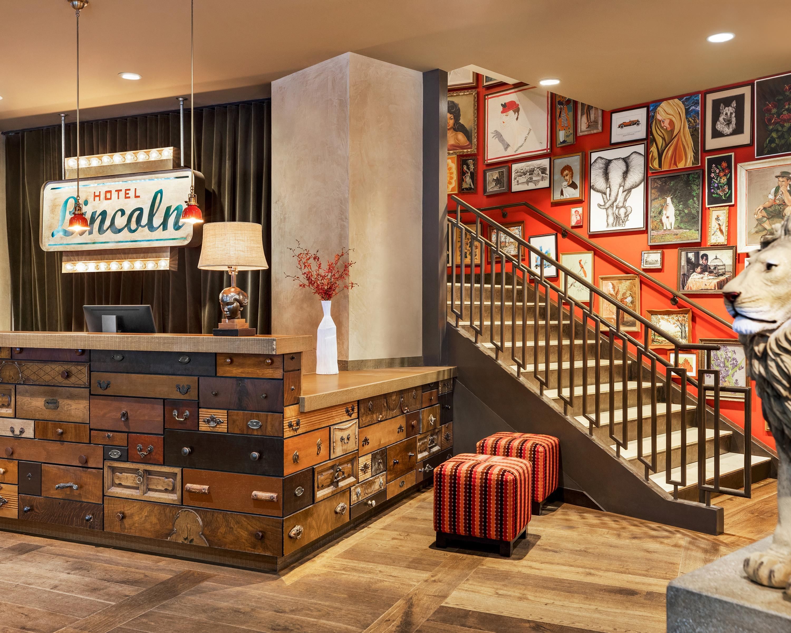 Um saguão de hotel com uma mesa de recepção, uma escadaria e uma parede decorada com diversas obras de arte.