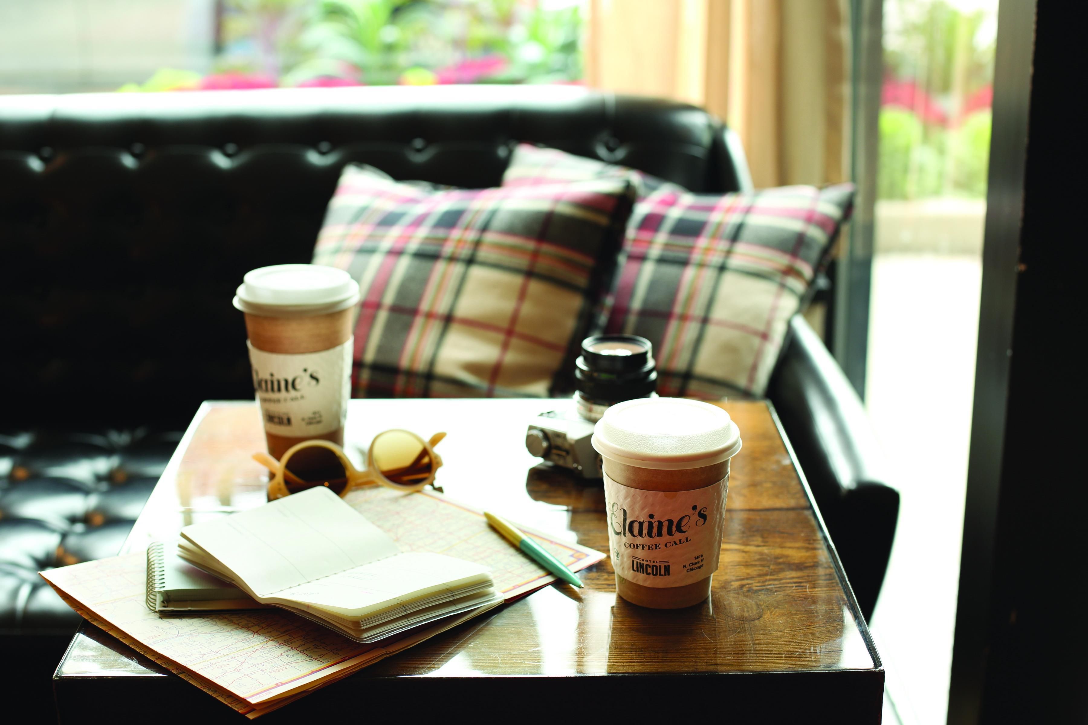A wooden table with a notebook, a pen, a pair of sunglasses, a camera, and two coffee cups from 'Laine's Coffee Call' on a leather couch with plaid cushions.