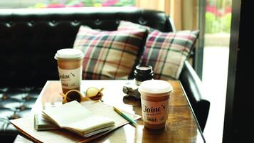 A wooden table with a notebook, a pen, a pair of sunglasses, a camera, and two coffee cups from 'Laine's Coffee Call' on a leather couch with plaid cushions.
