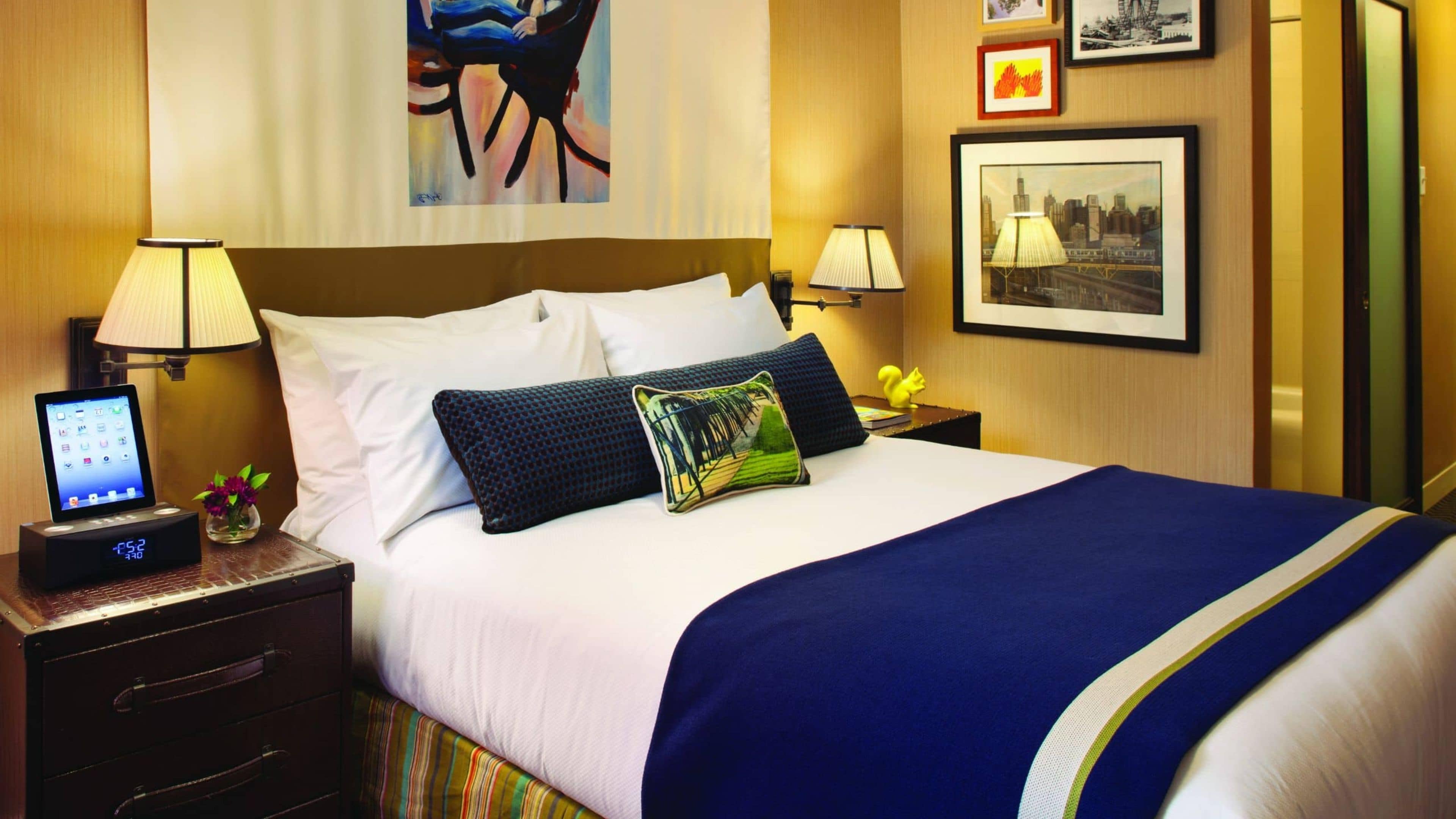 Habitación de hotel con cama, dos mesitas de noche, un reloj digital, una lámpara y obras de arte en las paredes.
