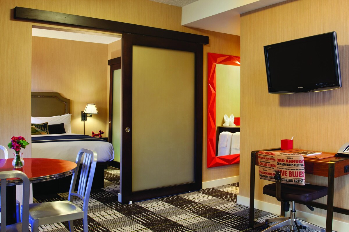 Habitación de hotel con cama, escritorio con silla, televisión y puerta que da a otra habitación.