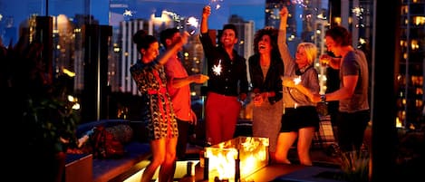 A group of people celebrating with sparklers on a rooftop terrace with a city skyline in the background.