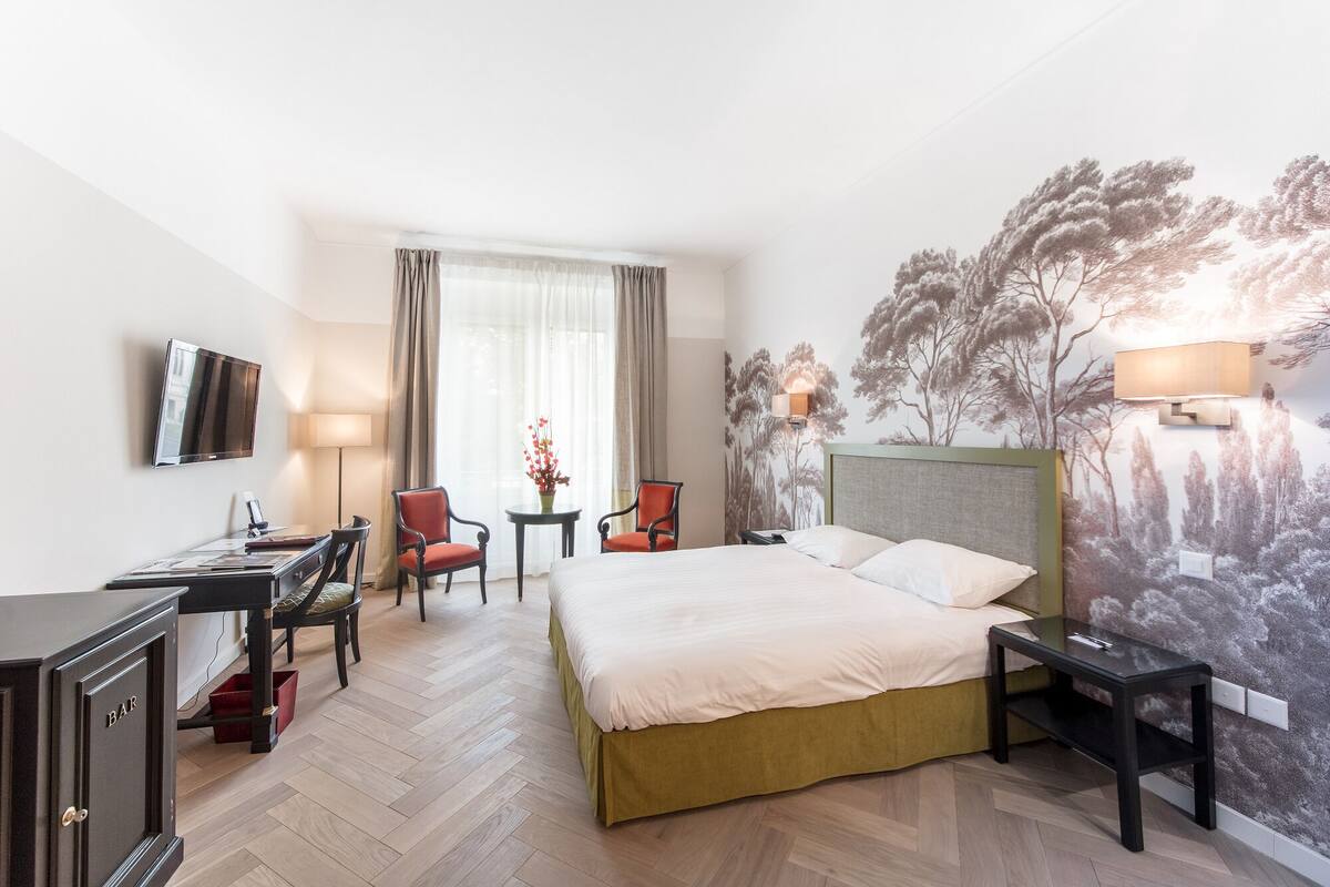 Habitación de hotel con cama, dos sillones, escritorio, televisión y un mural de un árbol en la pared.