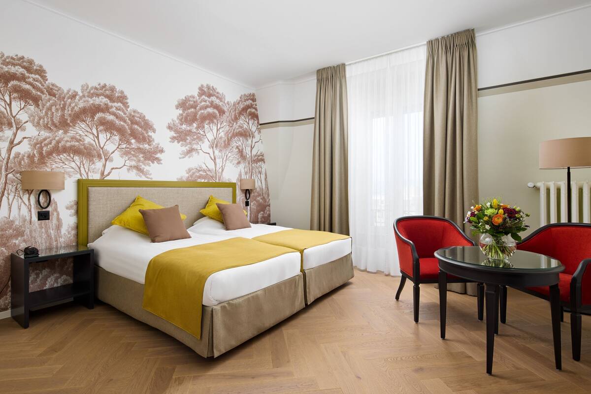 Una habitación de hotel con una cama, dos mesitas de noche, una lámpara de noche, una silla roja, una mesa redonda y un jarrón con flores.