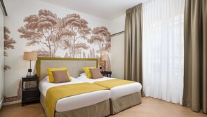 Een tweepersoonsbed met een beige en witte spreidekker, twee nachtkastjes met lampen, een muurschildering van bomen, en een raam met dunne gordijnen.