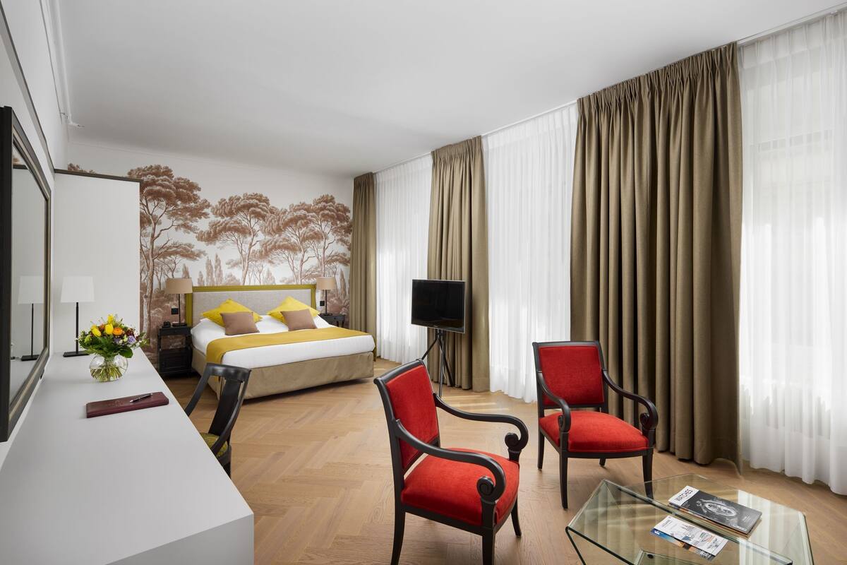 Una habitación de hotel con una cama, dos sillas rojas, una mesa de centro de vidrio, un televisor y un escritorio con un jarrón de flores.