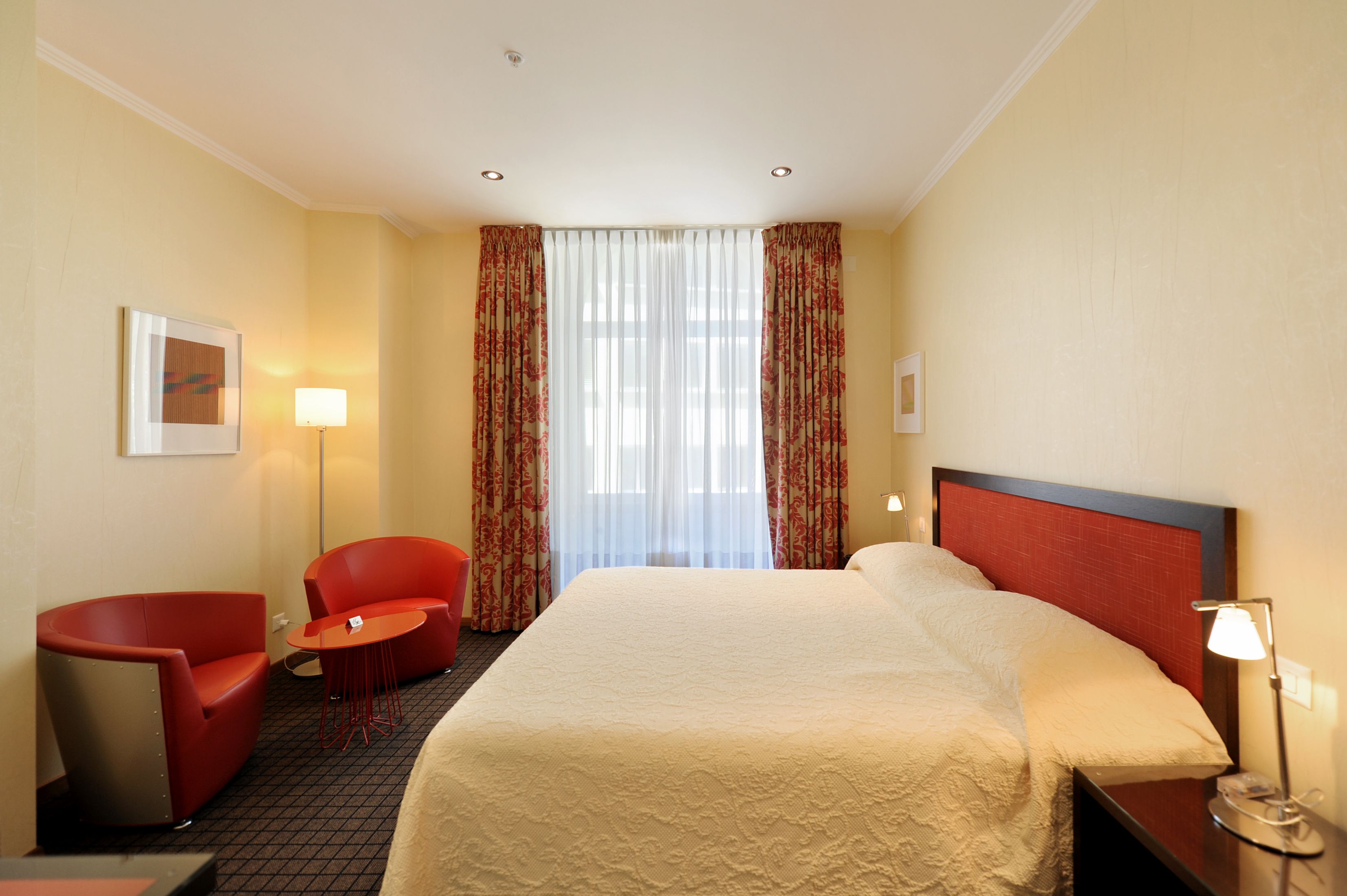 Une chambre d’hôtel avec un lit, un fauteuil rouge, une petite table, une lampe et une fenêtre avec des rideaux.