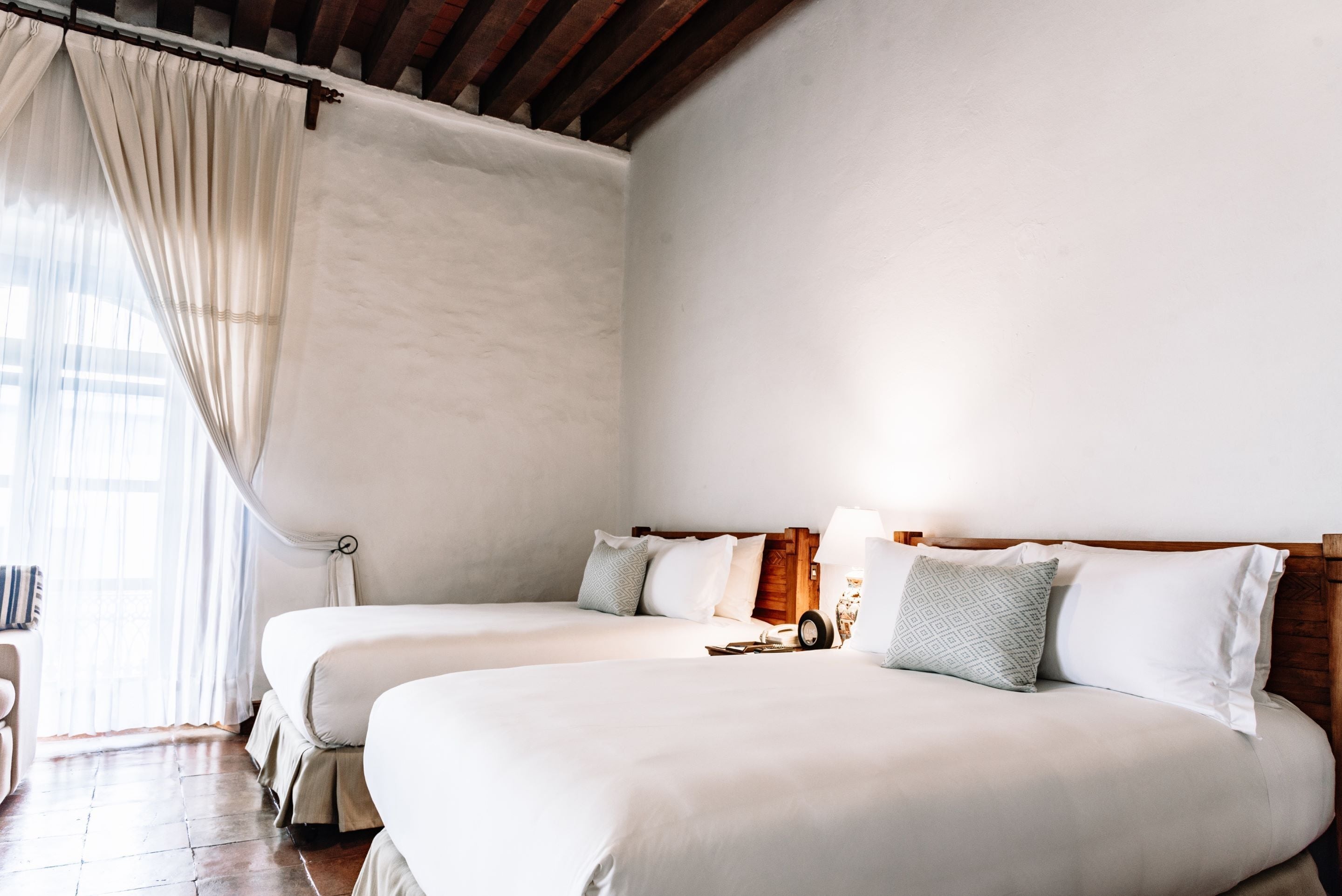 Habitación de hotel con dos camas, cabecera de madera, ventana con cortinas translúcidas y suelo de madera.