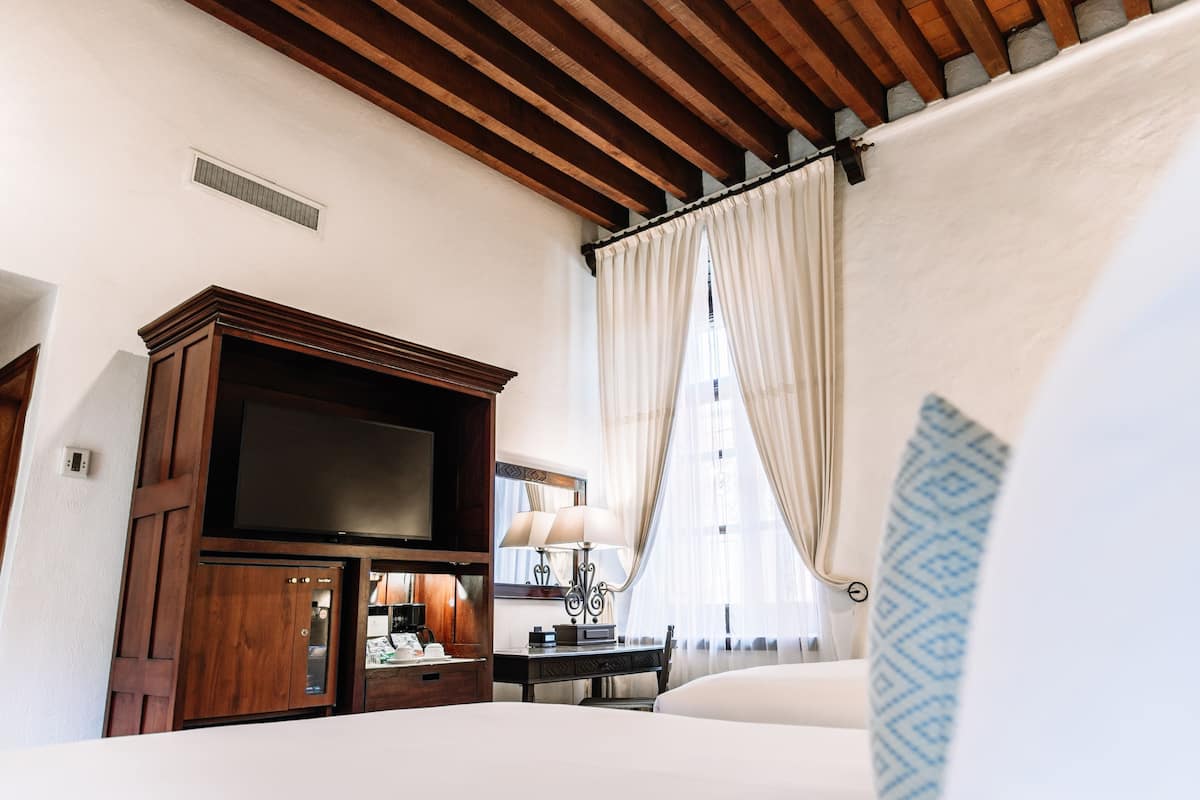 A hotel room with a wooden ceiling, a large flat-screen TV, a bedside table with a lamp, and a bed with white bedding.