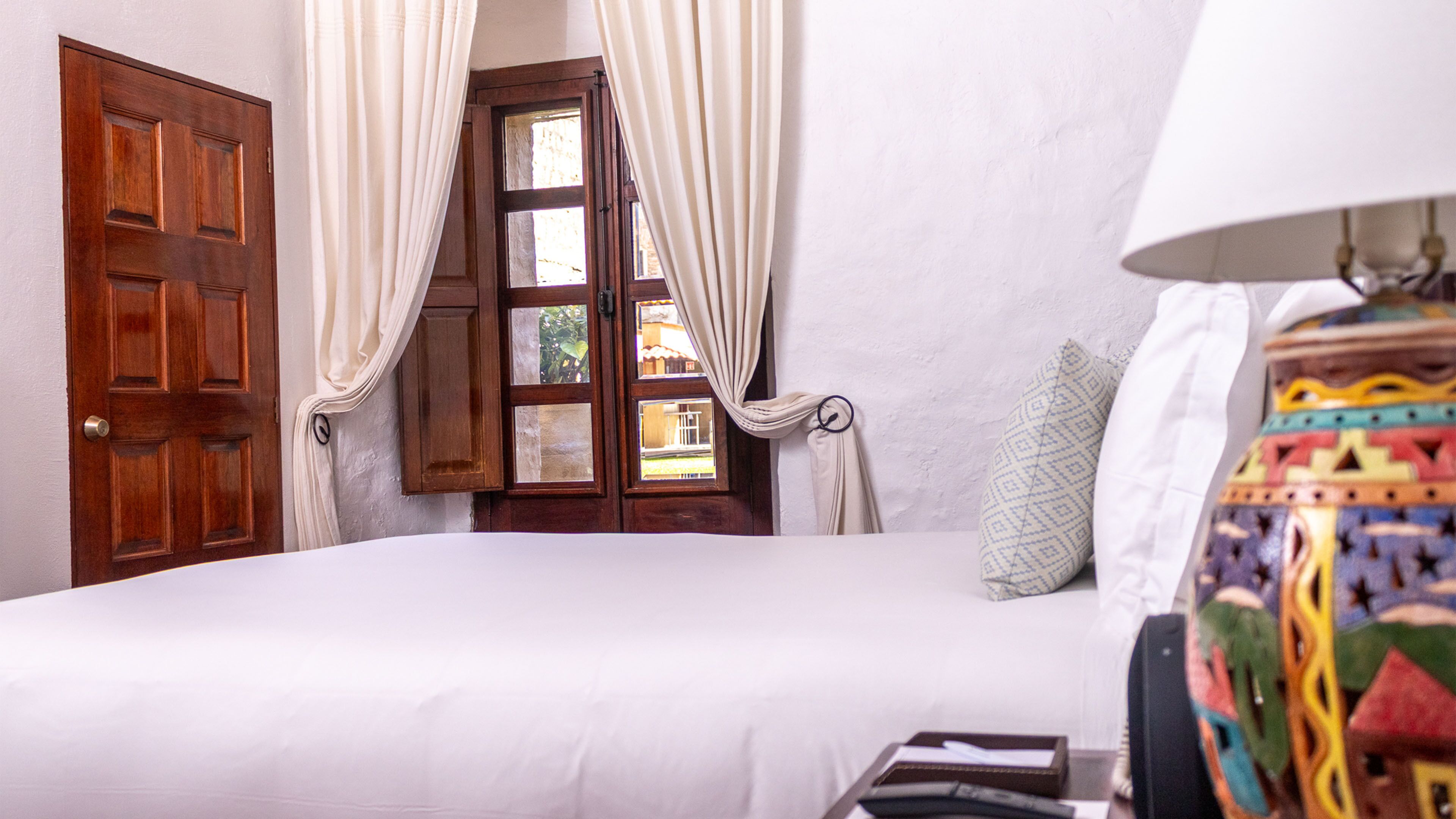 A bedroom with a bed, a window with curtains, a door, and a lamp.