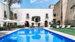 A swimming pool with lounge chairs and umbrellas, surrounded by a building with arched windows and a white facade.