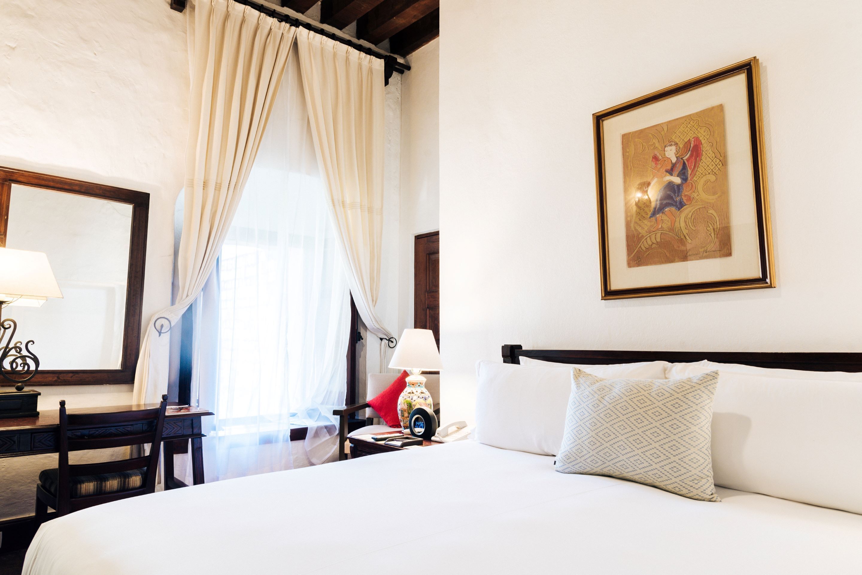 A hotel room with a bed, a desk, a chair, a lamp, a telephone, and a painting on the wall.