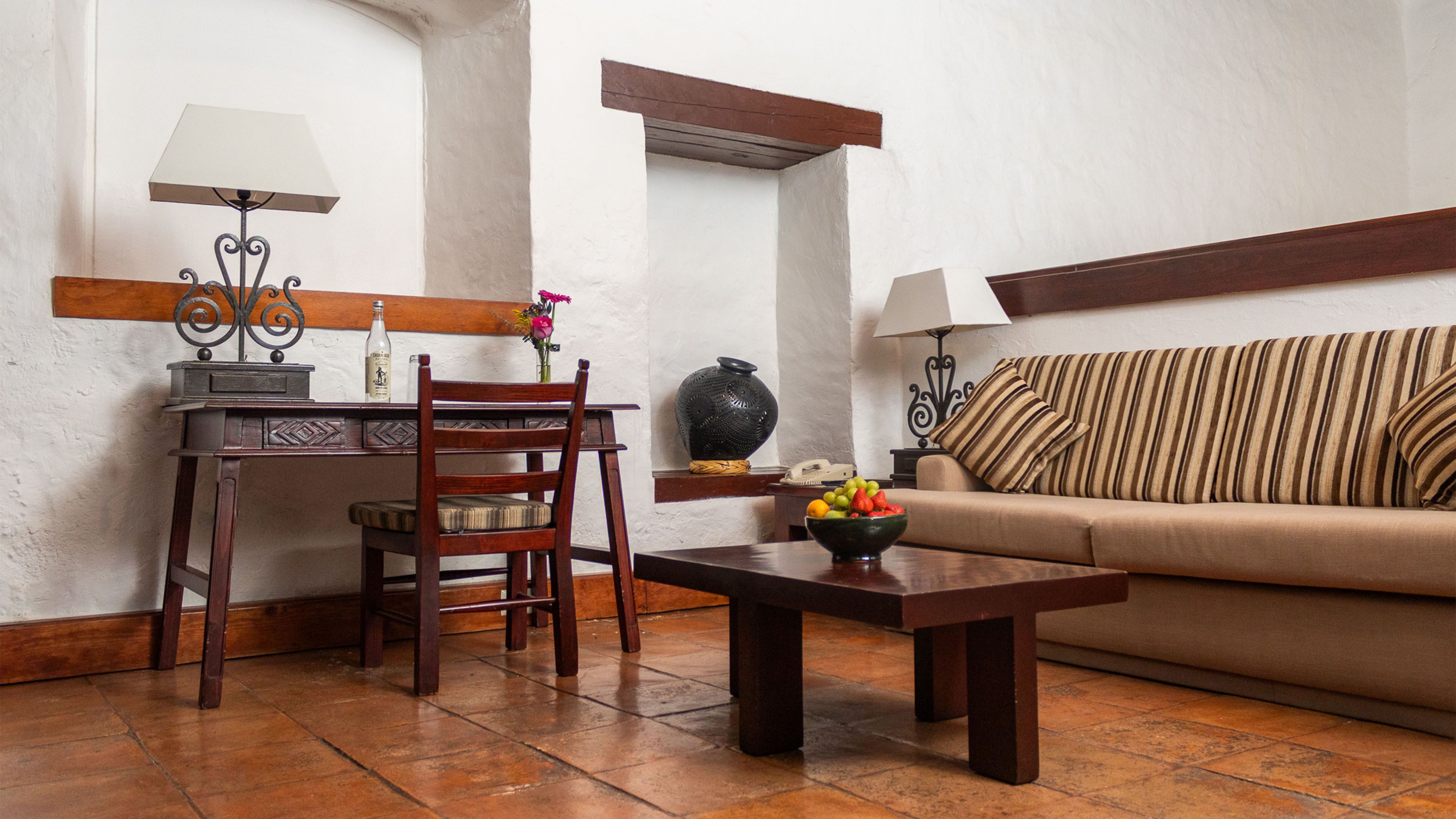 Une pièce comprenant une table en bois, une chaise, un canapé, une lampe, un vase et un saladier rempli de fruits.