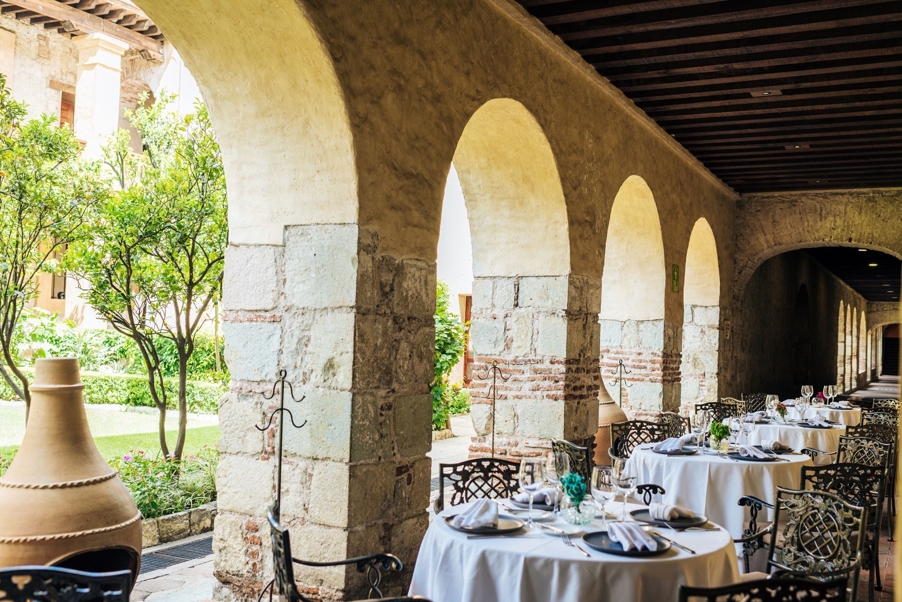 Un área de comedor al aire libre con mesas dispuestas para una comida, rodeada de arcos de piedra y vistas a un jardín.