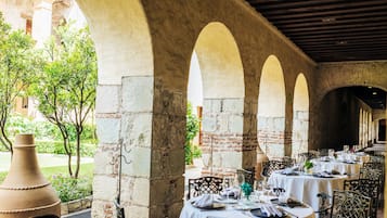 Un área de comedor al aire libre con mesas dispuestas para una comida, rodeada de arcos de piedra y vistas a un jardín.