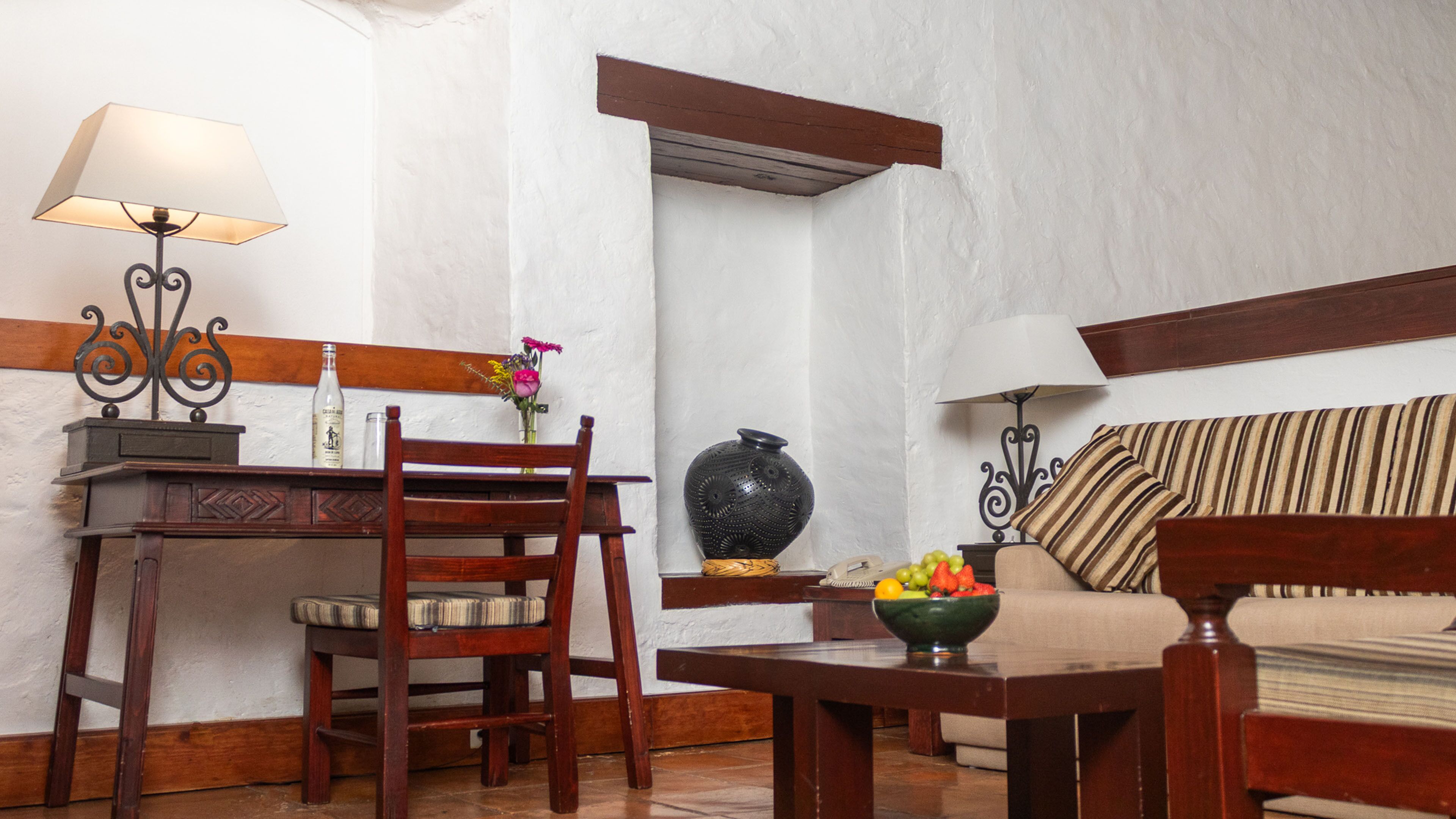 A room with a wooden chair, a small table with a bowl of fruit, a sofa, and a lamp.
