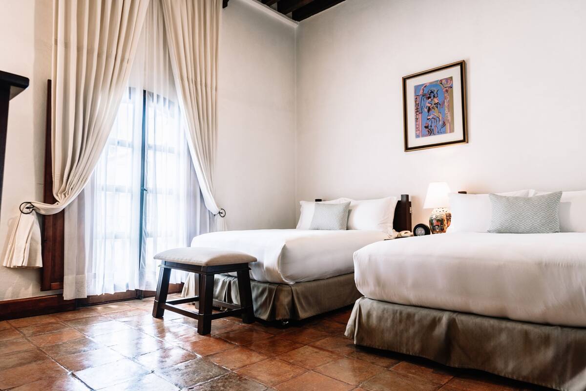 A hotel room with two beds, a bench, a window with curtains, and a framed picture on the wall.