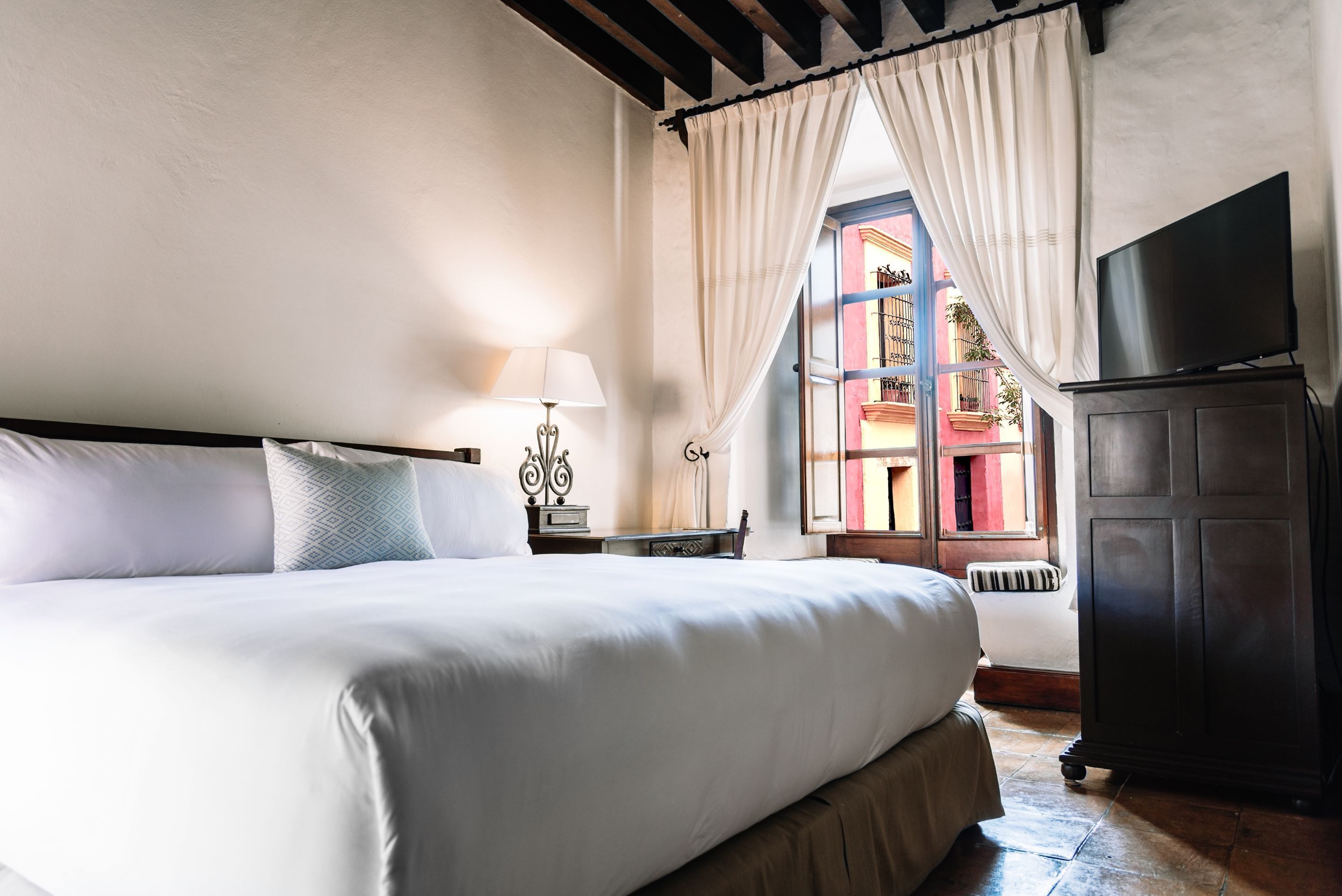 A neatly made bed with white linens, a bedside lamp, and a TV on a cabinet.