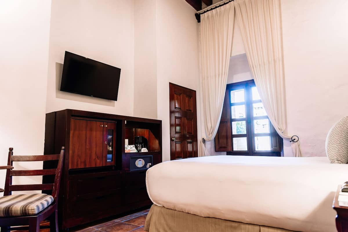 A bedroom with a bed, a television mounted on the wall, a wooden cabinet, a chair, and a window with curtains.