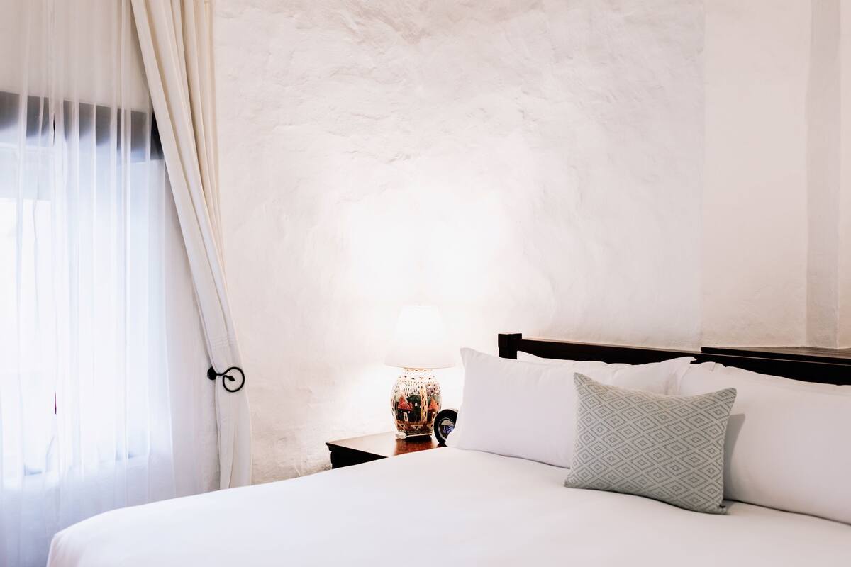 A hotel room with a bed, a nightstand, a lamp, and sheer curtains.