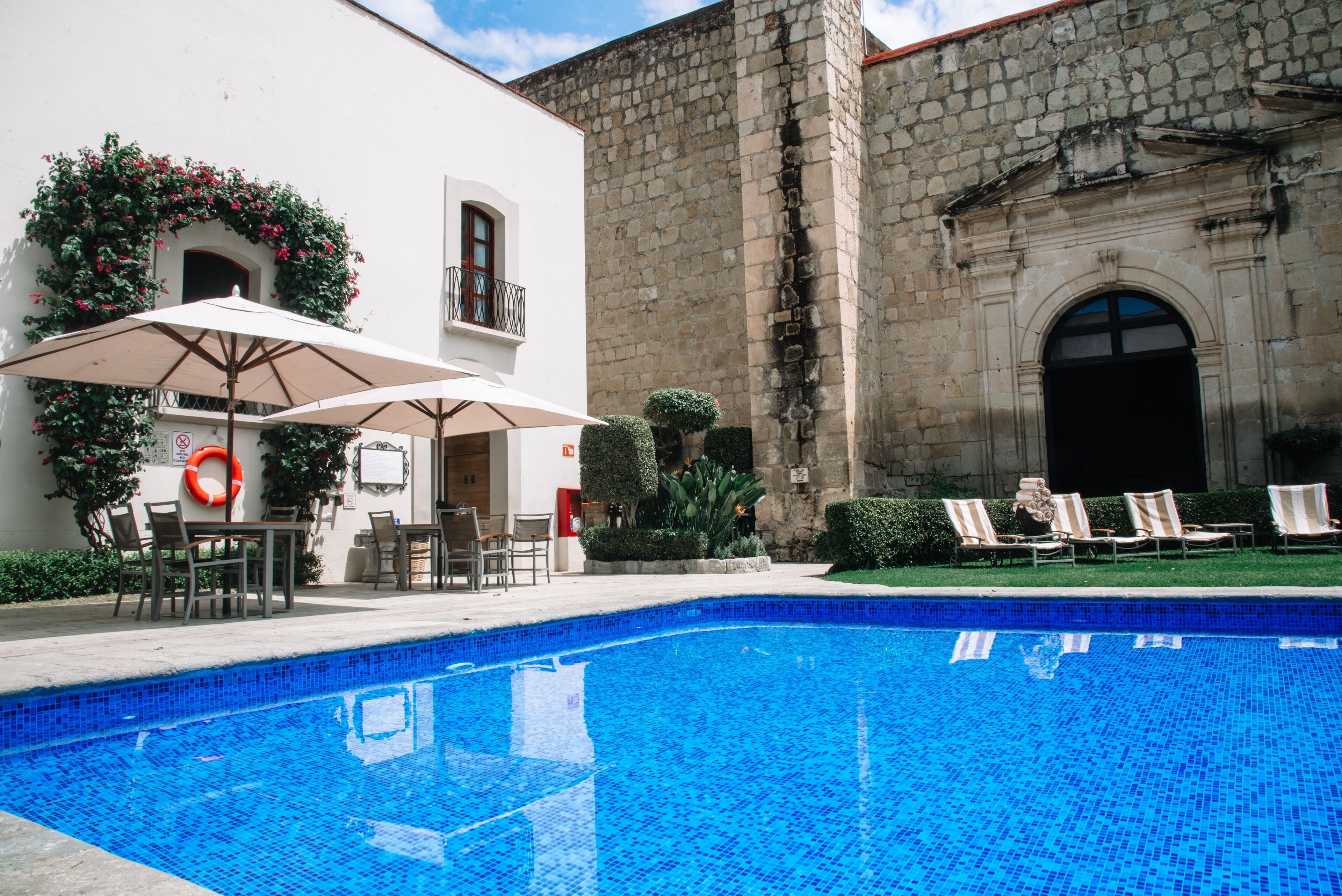 Una piscina con sillas de descanso y sombrillas, rodeada por un edificio de piedra con borde de azulejos.