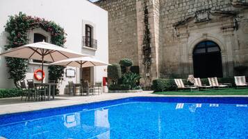 Una piscina con sillas de descanso y sombrillas, rodeada por un edificio de piedra con borde de azulejos.