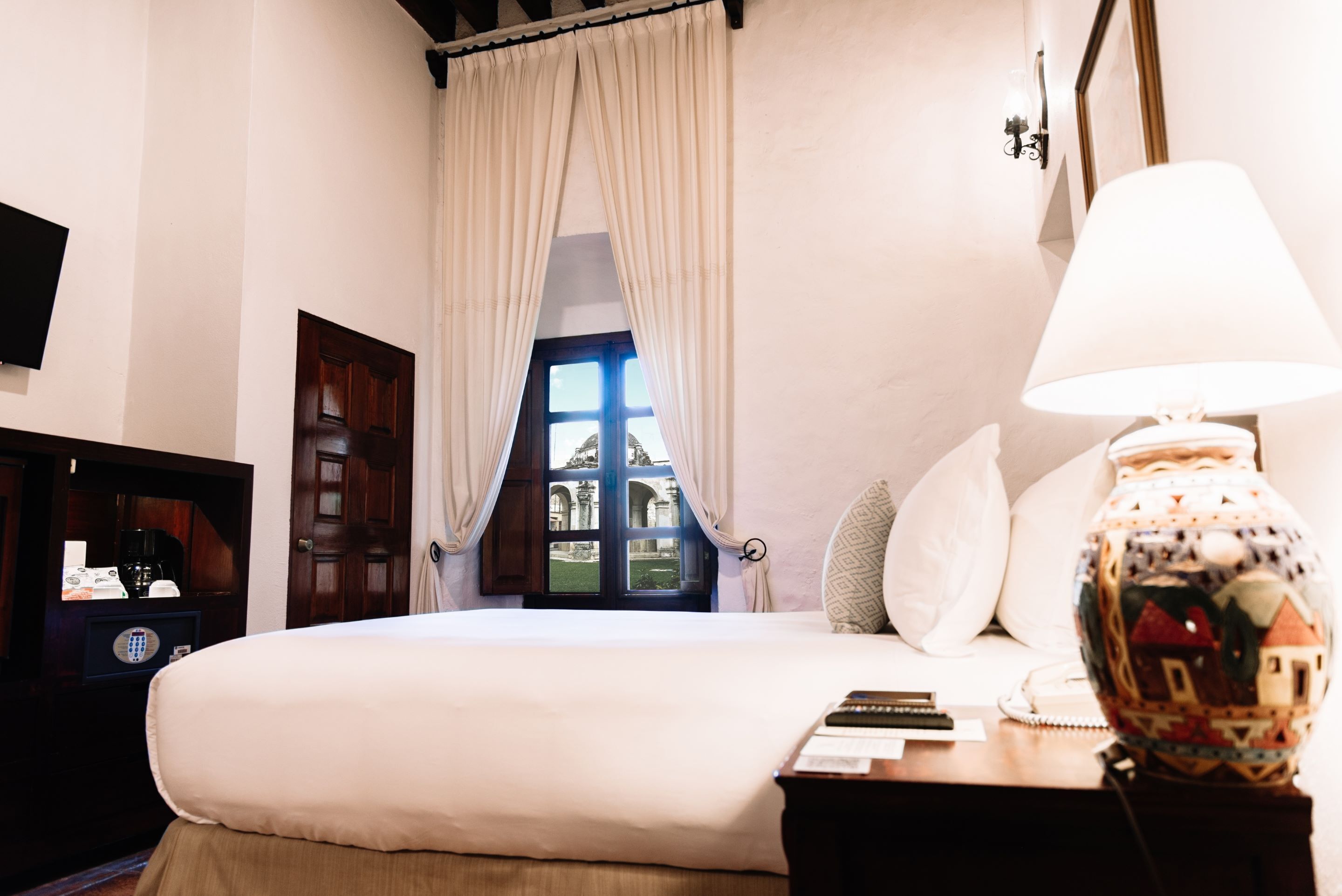 Habitación de hotel con una cama grande, una mesita de noche con lámpara, una ventana con cortinas y vistas a un edificio.
