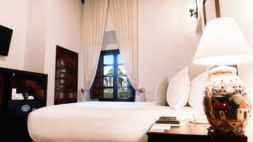 Habitación de hotel con una cama grande, una mesita de noche con lámpara, una ventana con cortinas y vistas a un edificio.