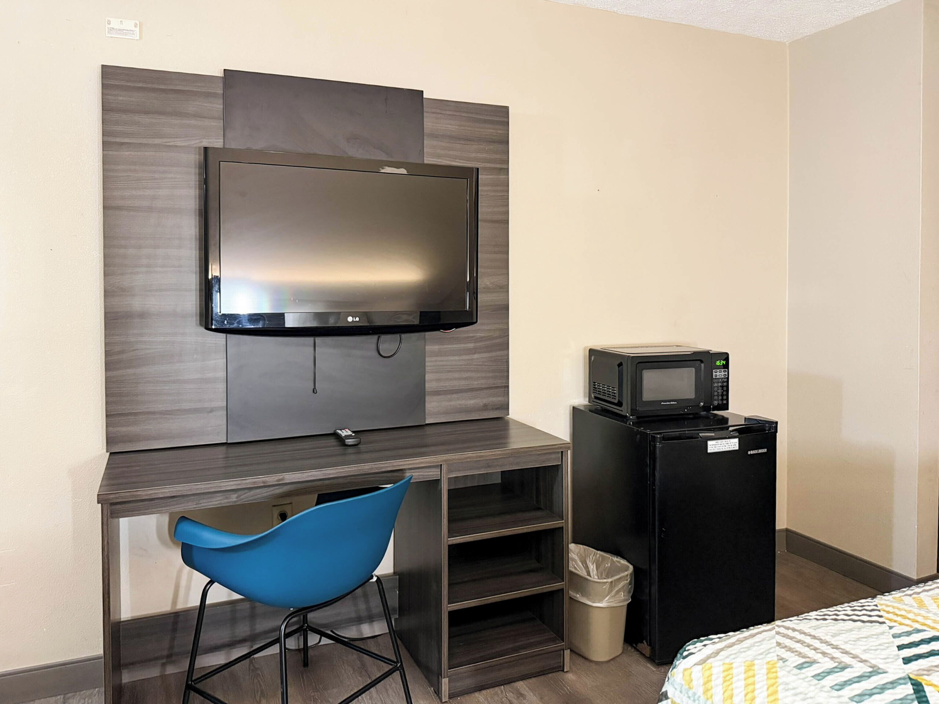 A hotel room with a flat-screen TV mounted on a wall, a small kitchenette with a microwave, a trash bin, and a blue chair.