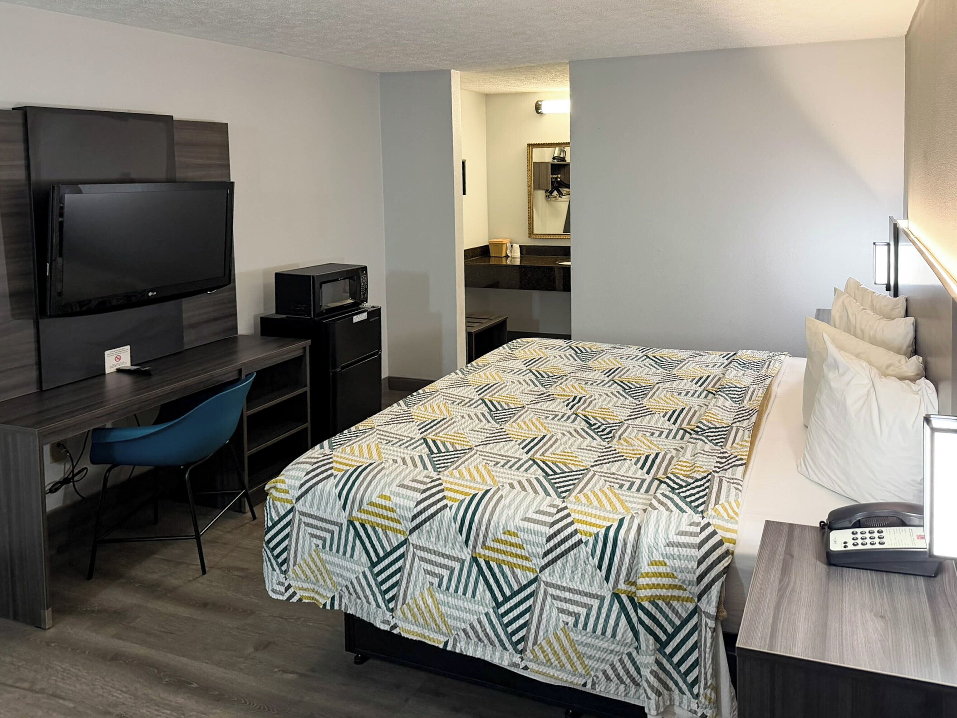 Habitación de hotel con cama, escritorio con televisor y microondas, una silla, mesita de noche con lámpara y espejo.