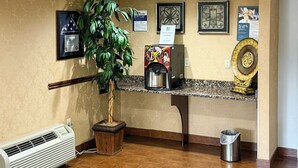 A reception area with a coffee machine, a trash can, a potted plant, and framed artwork on the wall. - Motel 6 Stafford, VA - Quantico (Stafford)