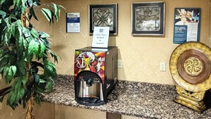 A coffee station with a coffee machine, a sign for coffee hours, framed artwork on the wall, and a decorative plate. - Motel 6 Stafford, VA - Quantico (Stafford)