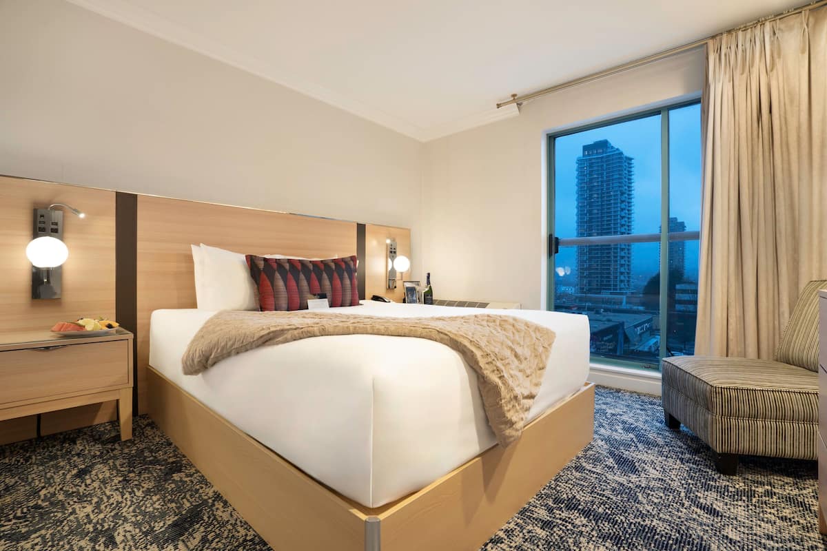 A hotel room with a large bed, a nightstand, a chair, a window with a city view, and a wall-mounted lamp.