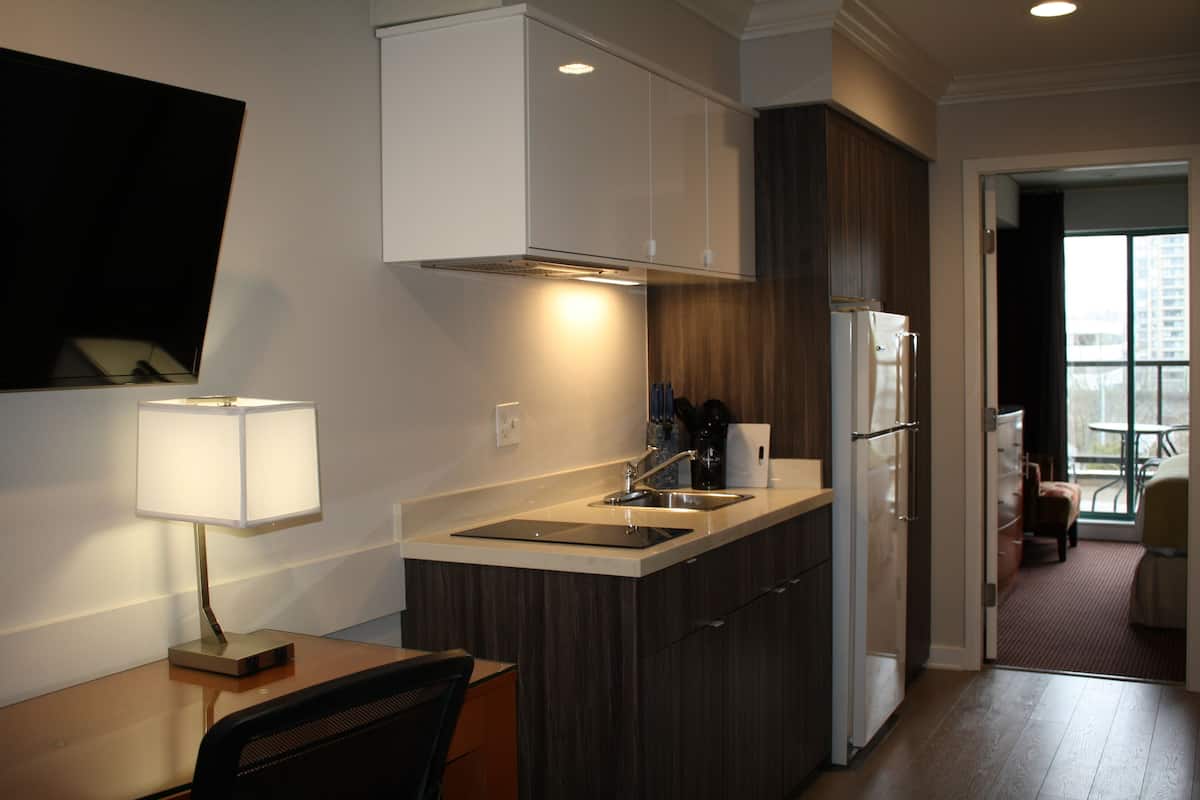 A compact kitchen with a sink, stove, and cabinets, a dining area with a chair, and a view of the outdoors through a door.