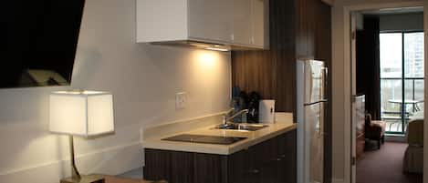 Cocina compacta con lavamanos, estufa y alacenas, zona de comedor con una silla, y vista al exterior a través de una puerta.