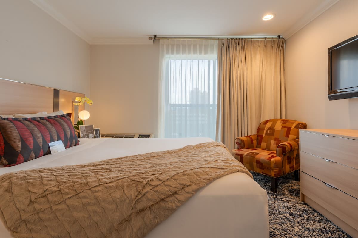 A hotel room with a large bed, a chair, a dresser, a flat-screen TV, and a window with curtains.