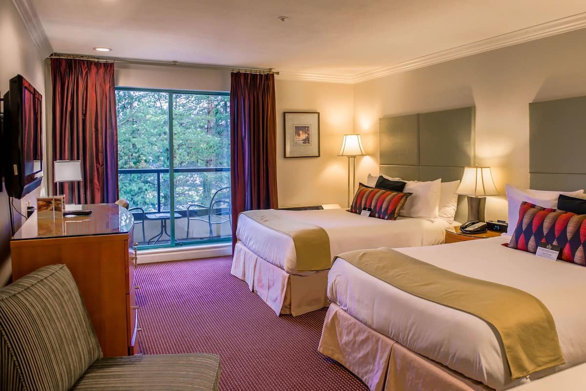 A hotel room with two beds, a desk with a chair, a TV, a window with curtains, and a view of trees.