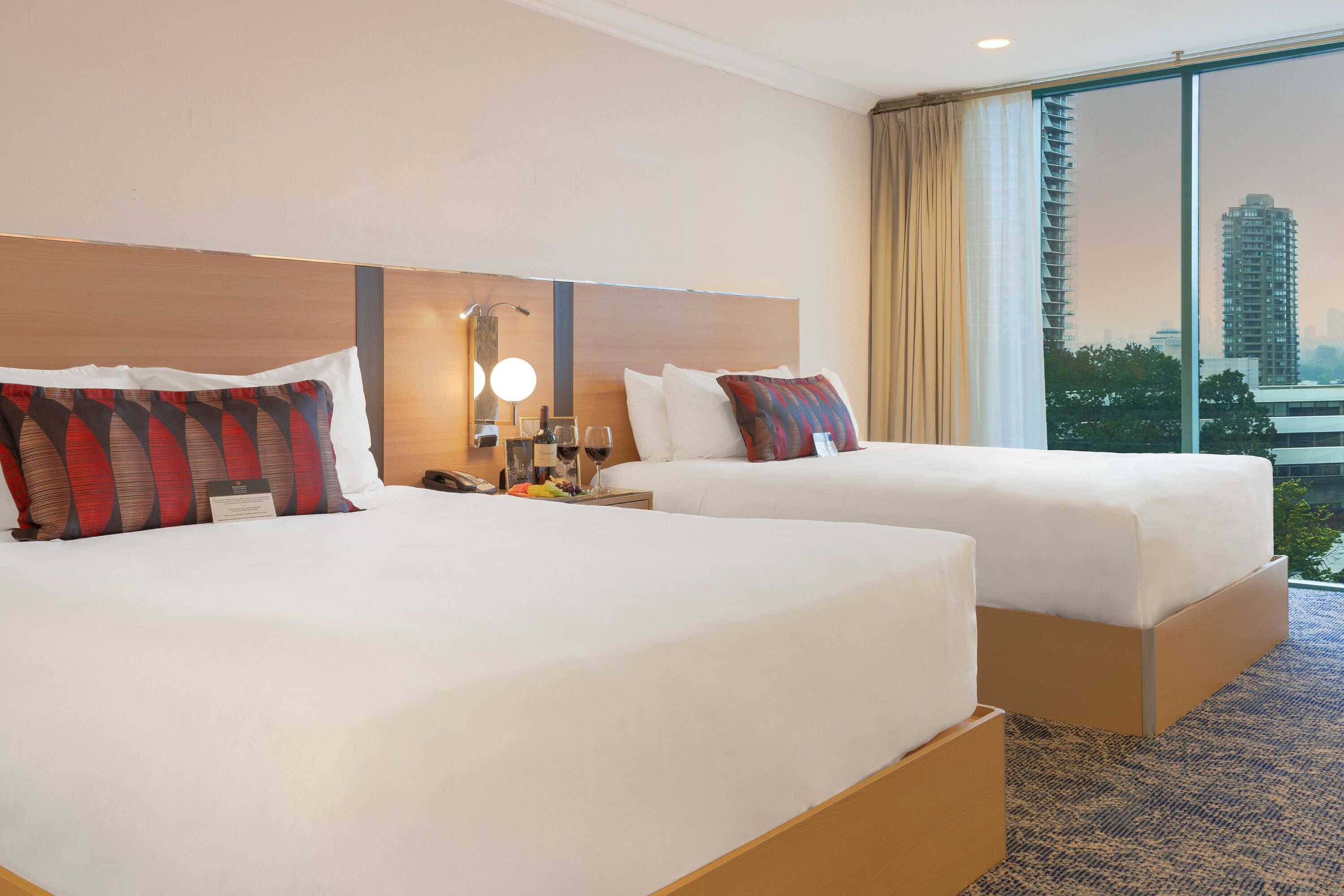 Een hotelkamer met twee bedden, een nachtkastje met een lamp, een telefoon en een uitzicht op een stadsbeeld door grote ramen.