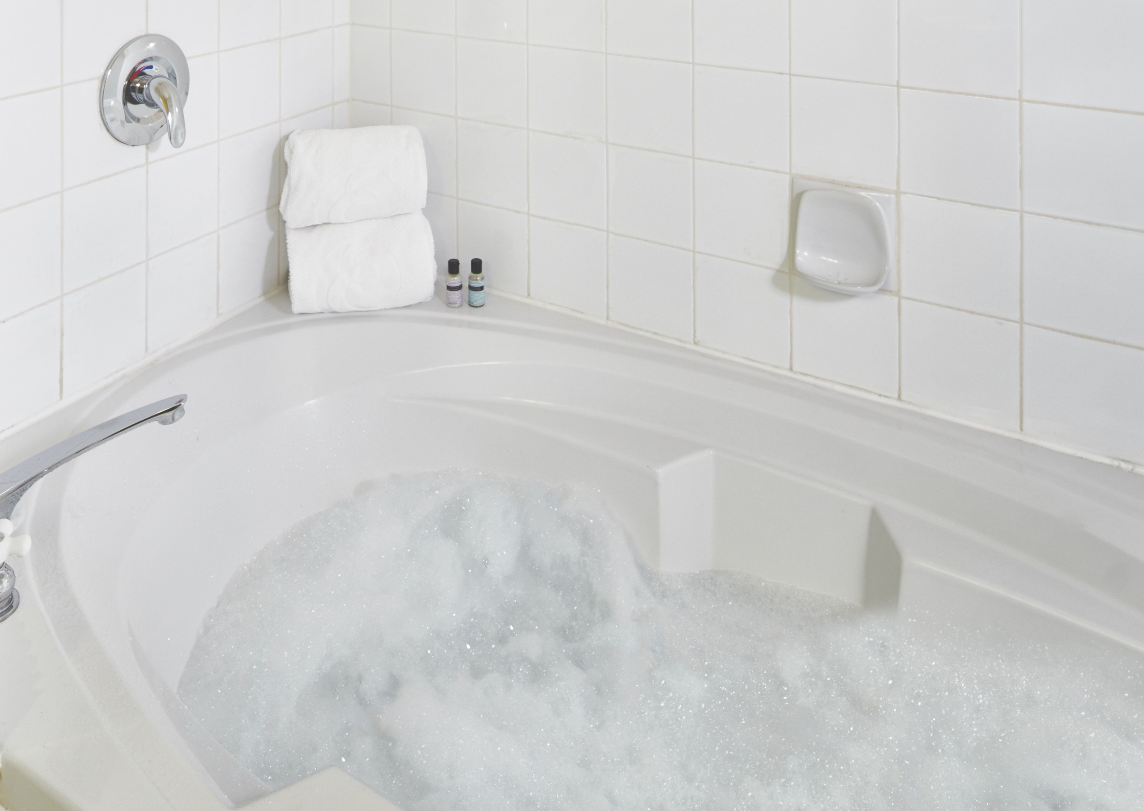 A bathtub filled with bubbles, a white towel, and a soap dispenser on a white tiled wall.