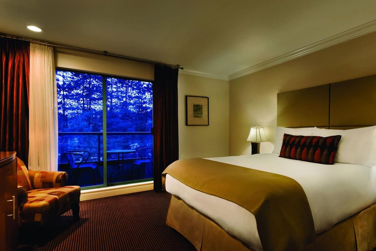 A hotel room with a large bed, a desk, a chair, a window with a view of trees, and a framed picture on the wall.