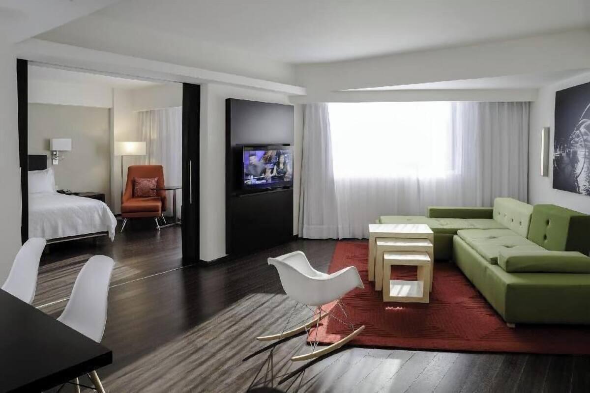 A hotel room with a bed, a TV, a green sofa, a white rocking chair, and a small table.