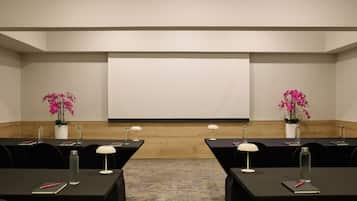 Un salón de conferencias con una pantalla de proyección grande, dos orquídeas rosadas en macetas y mesas con computadoras portátiles, cuadernos y botellas de agua.