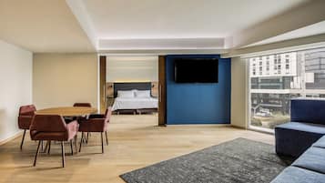 Una habitación de hotel moderna con zona de comedor, un ventanal con vista a la ciudad y un televisor de pantalla plana montado en la pared.