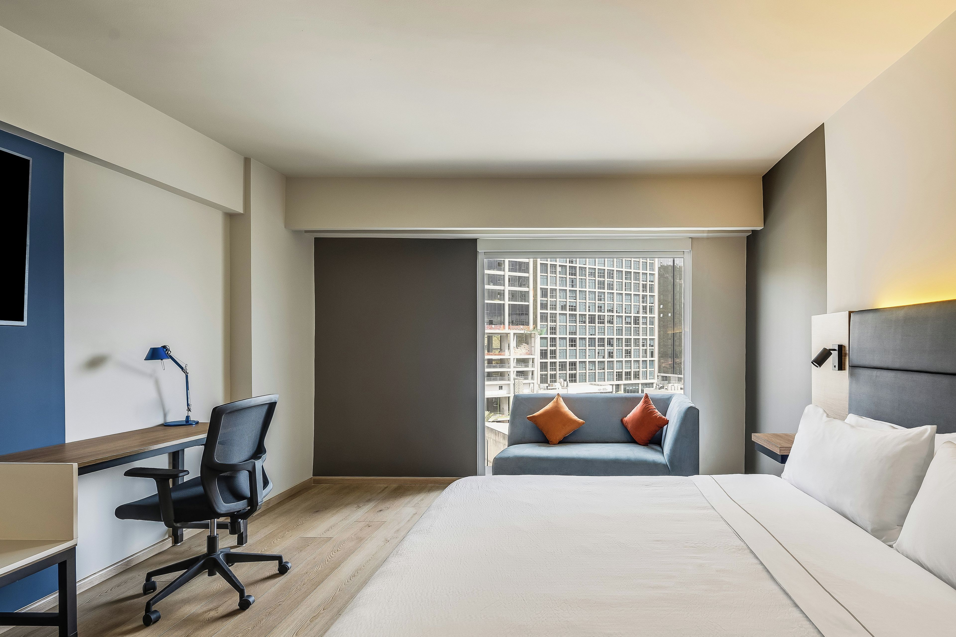Habitación de hotel con cama, escritorio, silla, ventana con vista a edificios y mesita de noche.