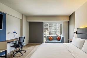 Habitación de hotel con cama, escritorio, silla, ventana con vista a edificios y mesita de noche.