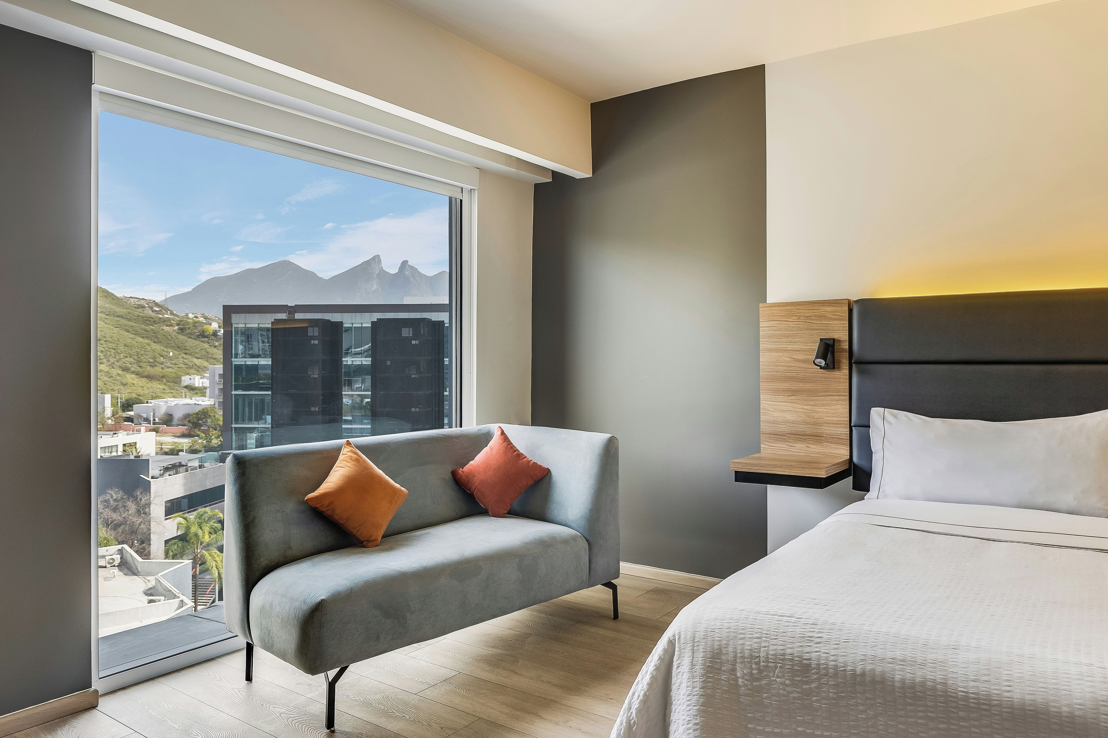 Una camera d'albergo moderna con una grande finestra, un divano grigio con due cuscini, un letto con biancheria bianca e un comodino in legno.