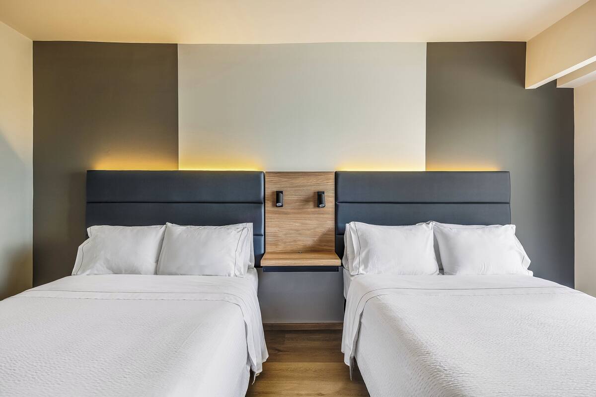 A hotel room with two beds, a wooden headboard, and a nightstand with a lamp.