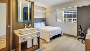 A modern bedroom with a large bed, a bedside table, a desk, and a window with a view. - Fiesta Inn Monterrey Valle (San Pedro Garza García)