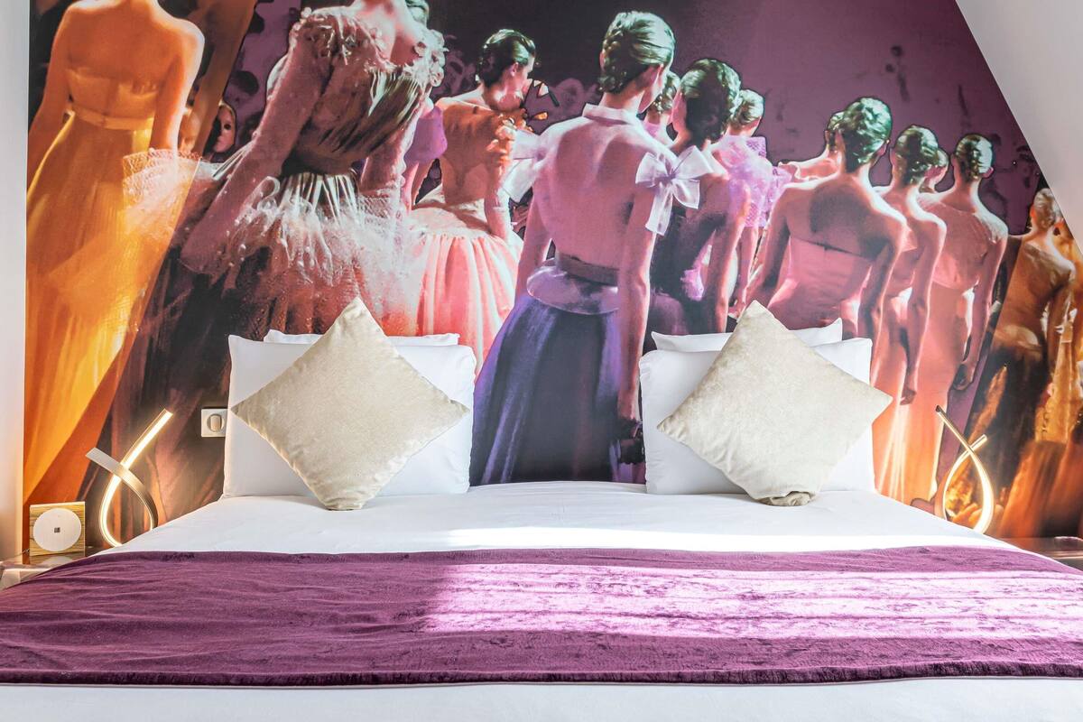 A bed with a purple blanket, white pillows, and a wall mural of people in dresses.